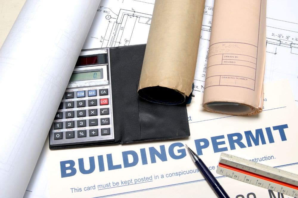 building permit and construction drawing under a calculator, triangular ruler, pen, and scrolled documents.