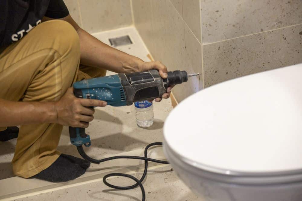 A bathroom remodeling contractor in khaki pants and a blue shirt uses a hand drill to drill holes through bathroom tiles, demonstrating how to choose a bathroom remodeling contractor with the right technical skills.