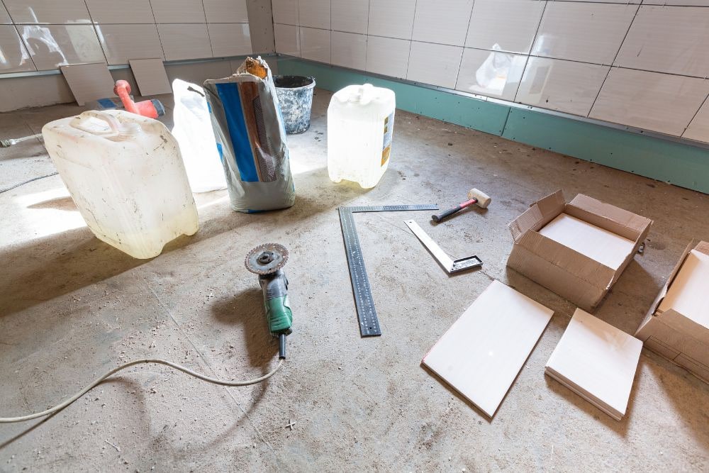 an unfinished bathroom floor with a bag of cement, water containers, newly opened boxes of tiles, and tools including a tile cutter, framing squares, and mallet; the image implies an ongoing bathroom remodel construction