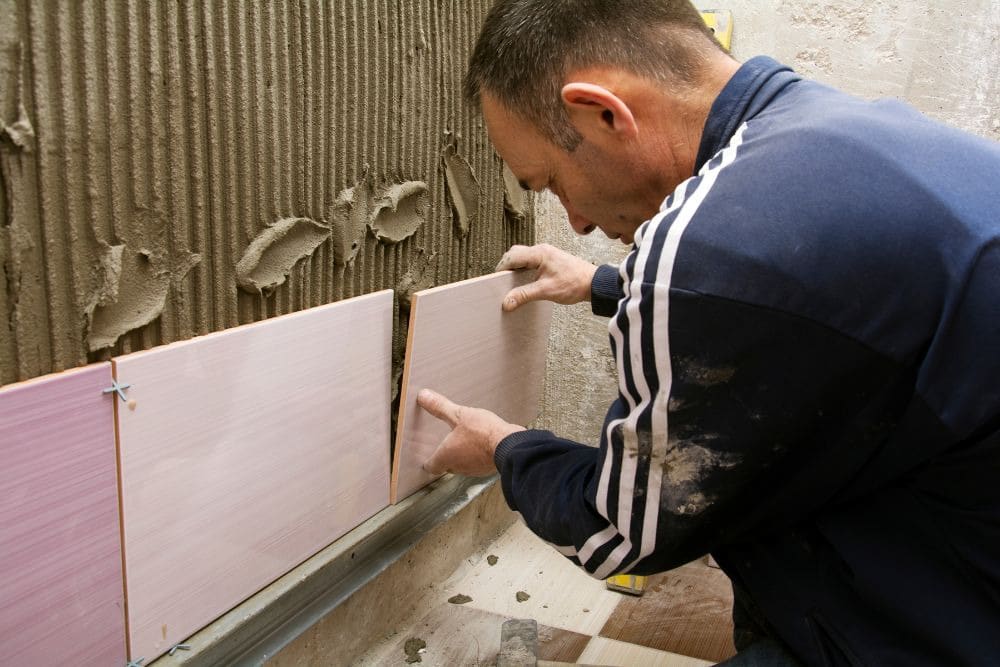 a male contractor wearing a blue tracksuit carefully installs bathroom wall tiles, representing the technical expertise to look for when preparing questions to ask a bathroom remodeling contractor.